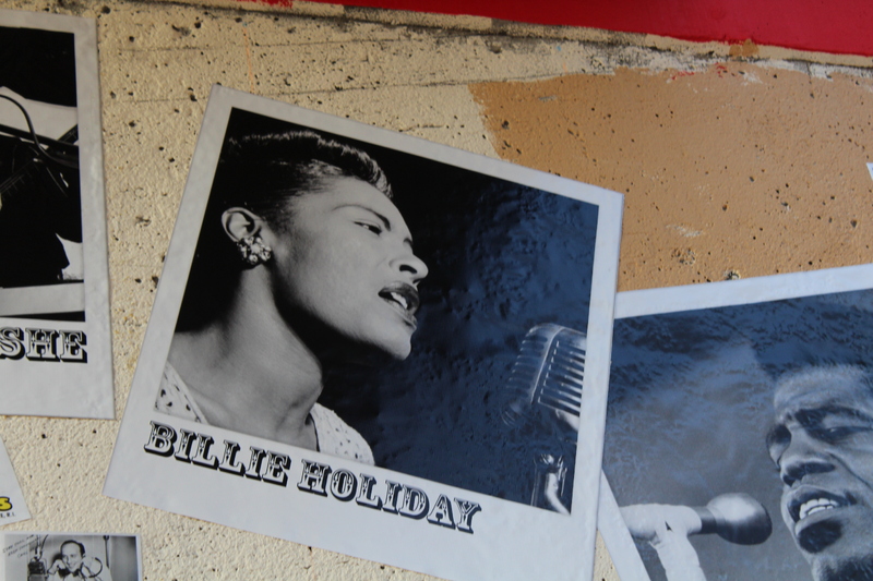 Black-and-white photo of labeled Billie Holiday on textured wall, showing singer mid-performance at a vintage microphone.